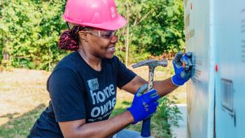 A woman in a pink hardhat and homeowner T-shirt smiles as she hammers.